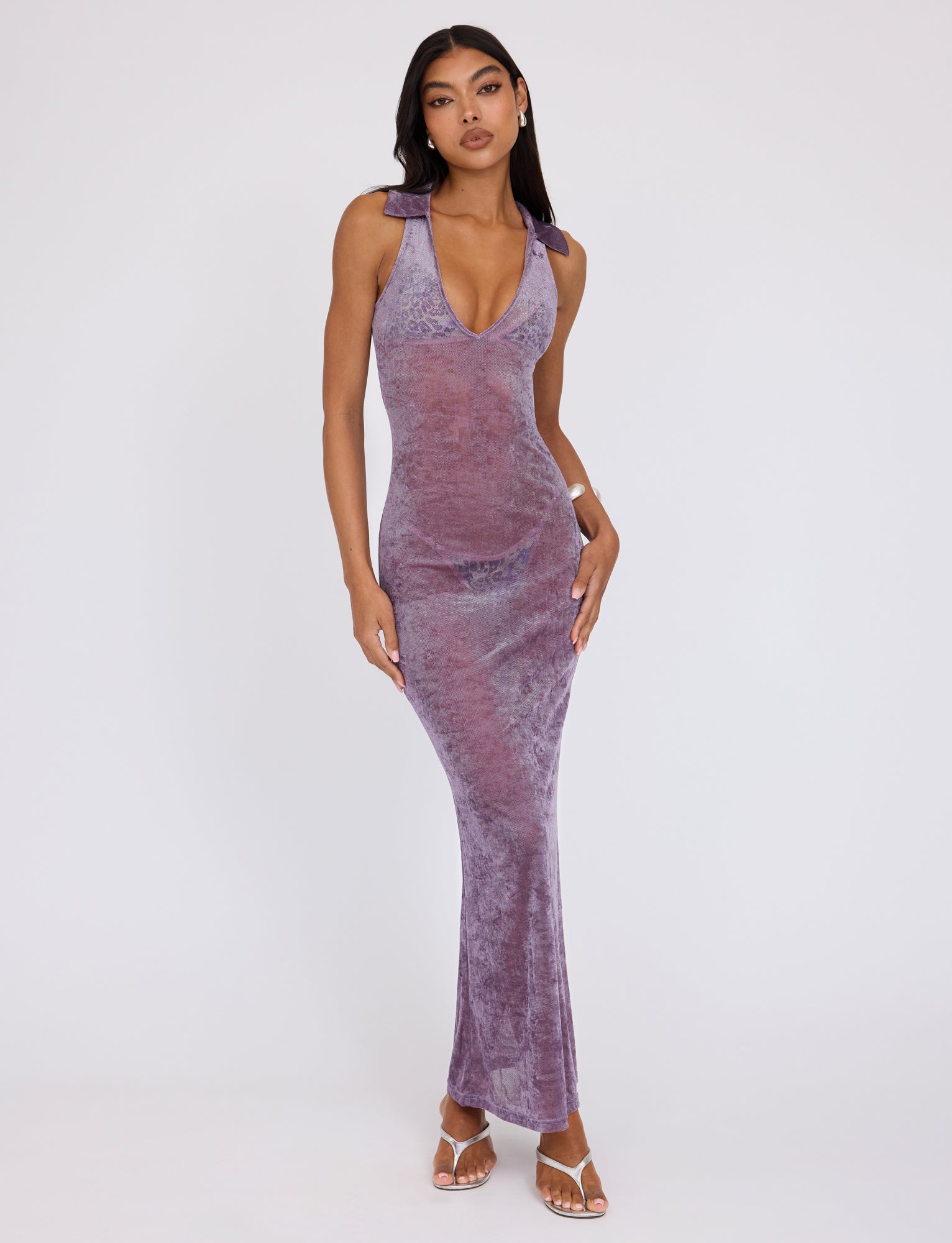 A woman stands against a plain white background, wearing the sleeveless, V-neck Abril Dress - Purple from TIGERMIST. She has long, dark hair and is sporting white open-toe heels. Her right hand rests on her hip while her left hand lightly touches her thigh.