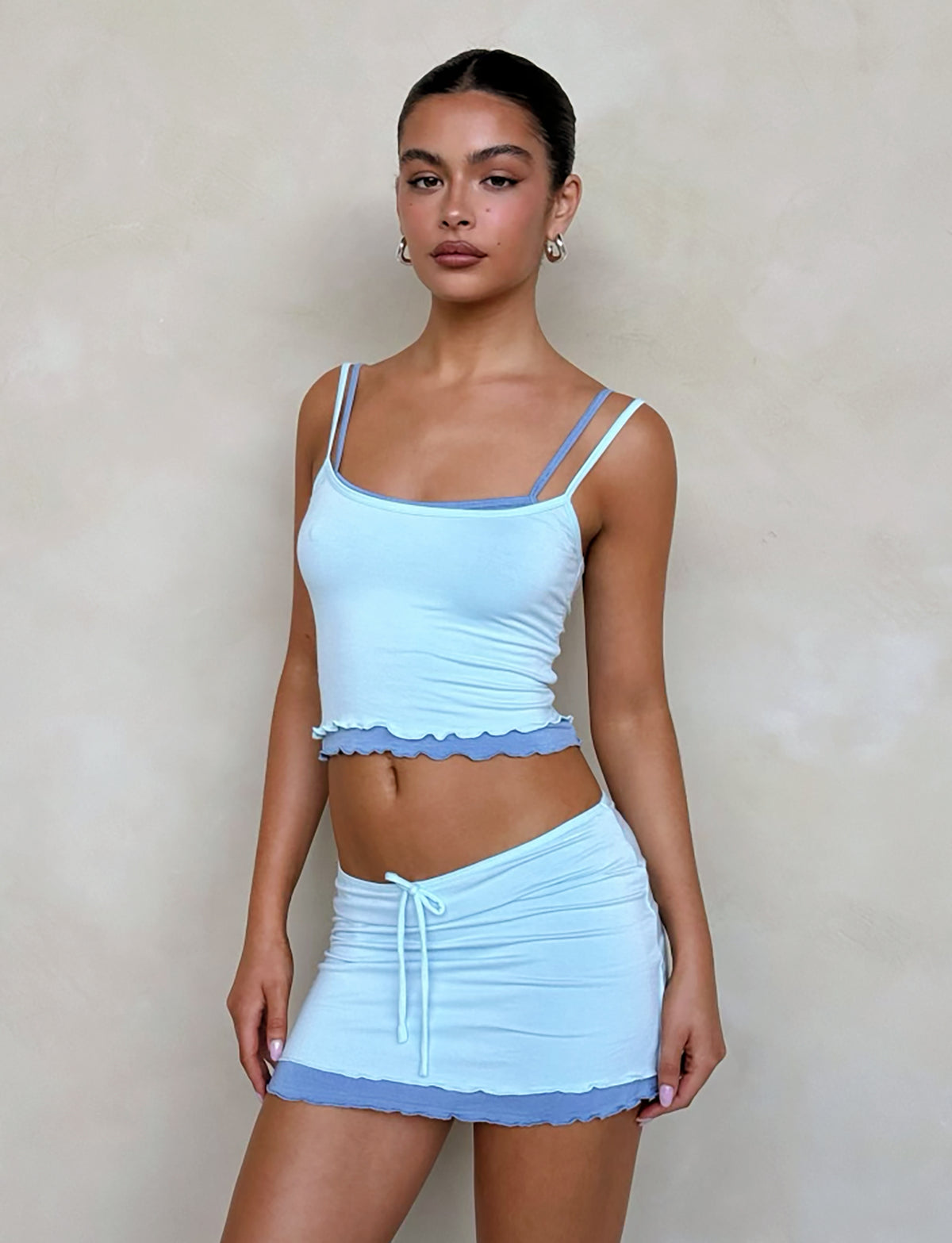 A woman stands against a plain background wearing the ADAIR TOP - BLUE from TIGERMIST, a light blue spaghetti strap top paired with a matching mini skirt that features ruffled edges. She has her hair pulled back, is wearing earrings, and looks confidently at the camera.
