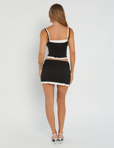 A woman with long hair is standing and facing away from the camera, showcasing her chic black top with thin straps from TIGERMIST. She pairs it perfectly with the ADAIR SKIRT - BLACK by TIGERMIST, which features a white hem. Completing the look are stylish white high-heeled shoes, all set against a plain white wall backdrop.
