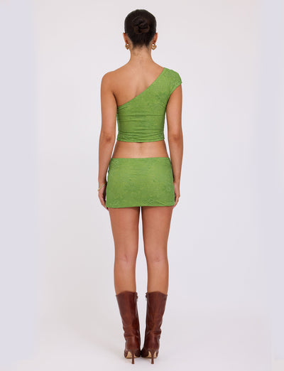 A person with their back to the camera is wearing the AANYA DRESS - GREEN LACE from TIGERMIST, with its textured, green, one-shoulder design and lace overlay. They have their hair styled in a bun and are accessorized with brown knee-high boots, set against a plain white background.