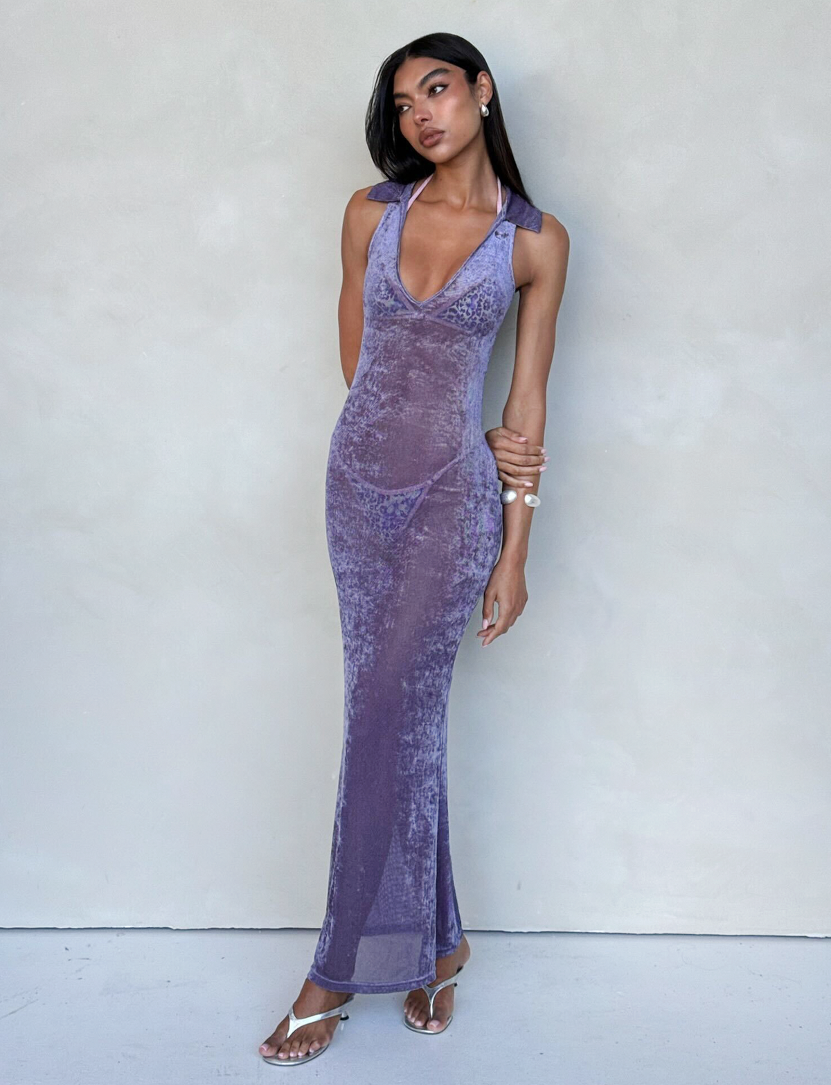 A woman with long hair stands against a pale background, wearing the sleeveless, floor-length, form-fitting ABRIL DRESS - PURPLE from TIGERMIST, featuring a deep neckline. She has one hand on her hip and the other resting against her opposite arm, accessorized with stylish sandals and bracelets.