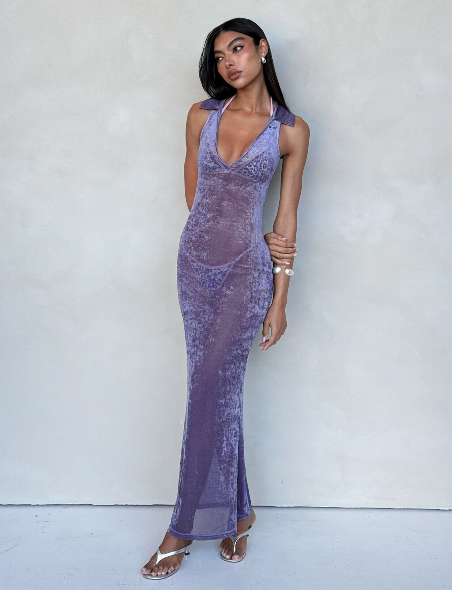A woman with long hair stands against a pale background, wearing the sleeveless, floor-length, form-fitting ABRIL DRESS - PURPLE from TIGERMIST, featuring a deep neckline. She has one hand on her hip and the other resting against her opposite arm, accessorized with stylish sandals and bracelets.