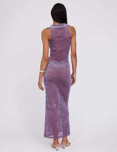 An individual with long, dark hair is standing with their back to the camera, donning a sleeveless, form-fitting, ankle-length ABRIL DRESS - PURPLE from TIGERMIST made from a sheer, shimmery fabric. They are also wearing white sandals and a white bracelet on their right wrist.