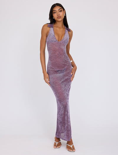 A woman stands against a plain white background, wearing the sleeveless, V-neck Abril Dress - Purple from TIGERMIST. She has long, dark hair and is sporting white open-toe heels. Her right hand rests on her hip while her left hand lightly touches her thigh.